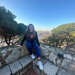 Young woman sits on a stone ledge with tress and city rooftops behind her.