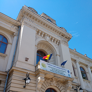 The front of a pale stone building with two flags waving and a blue sky in the background.