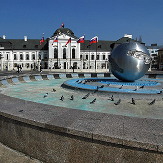 A fountain with a silver globe sits in front of a white building with a row of Slovakian flags.