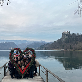 College students pose peeking through a large heart-shaped wreath on a dock with a lake and castle in the background.