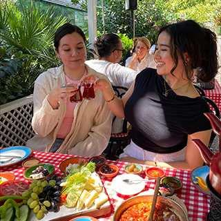 Two young woman clink their tea glasses at a cafe with a spread of fruit and cheese on the table in front of them.