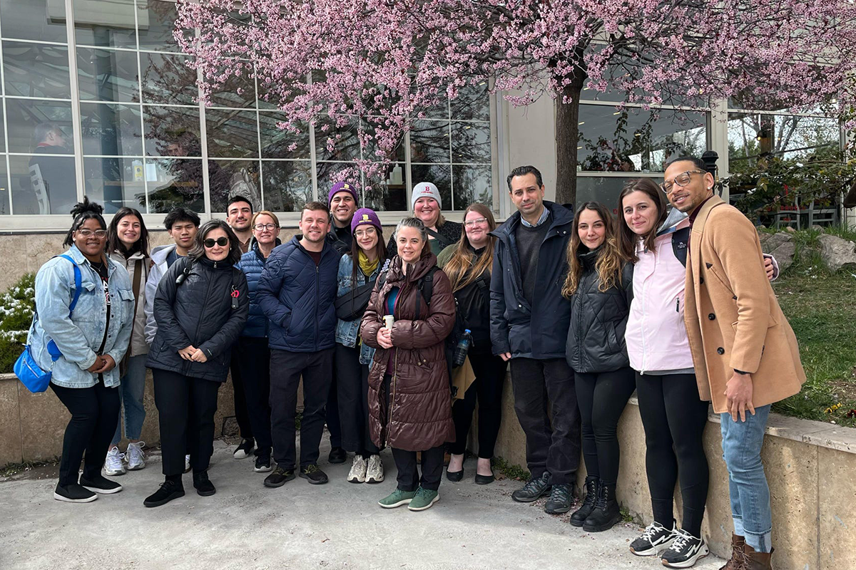 A large group of adults pose outside a restaurant with the branches of a pink flowered tree above.