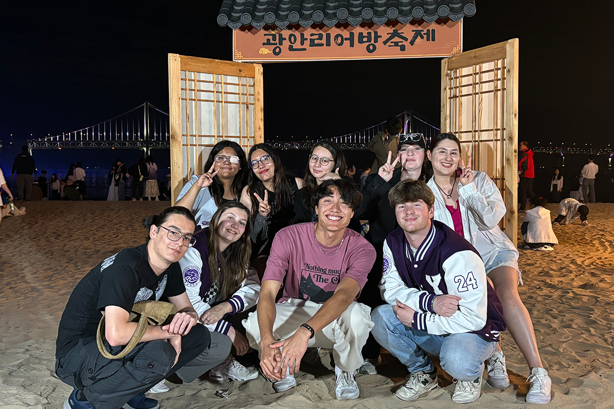 Group of college students pose together on a beach at night with a wooden structure in the background.