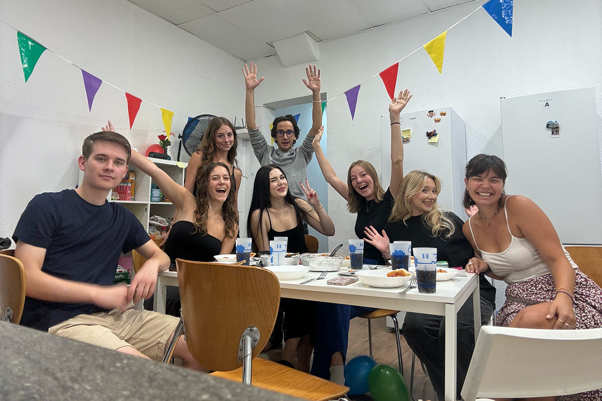 College students pose together around a dining table with food and drinks.