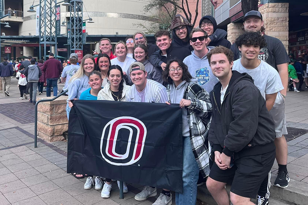 UNO students pose with the UNO flag at a baseball stadium.