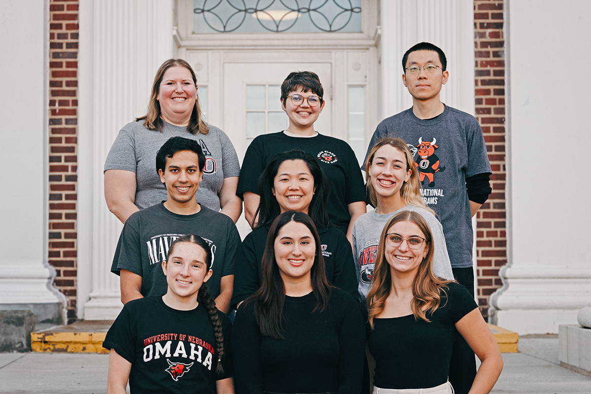 Group of 9 people posing in front of a building.