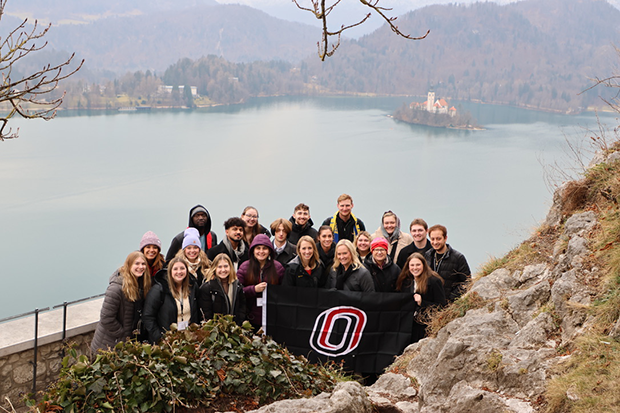 Group of students pose with the UNO flag in front of a lake in Slovenia.