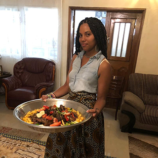 Young woman stands in a living room holding a large silver bowl full or yellow rice, meat, and vegetables.