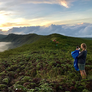 Young woman standing on a lush hill takes a photo of the beach in the distance.