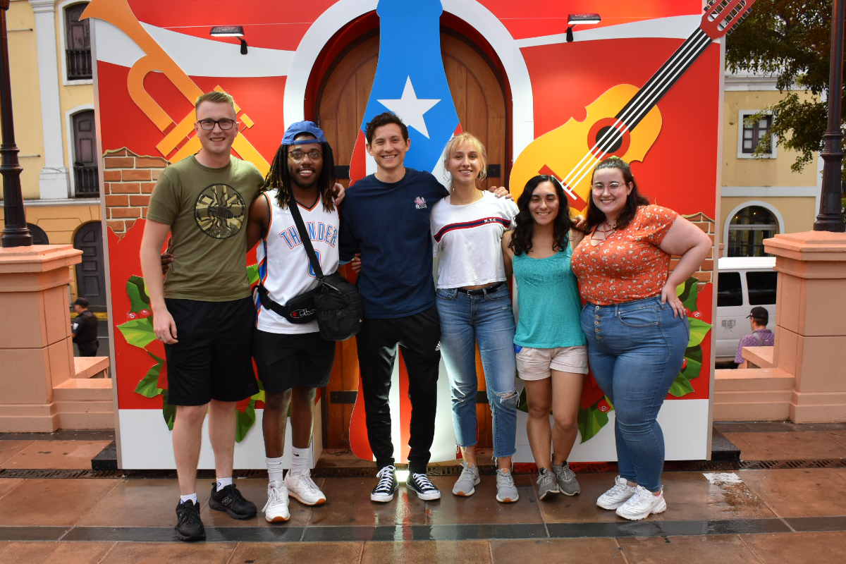 A group of 6 students pose in front of a colorful music themed backdrop outside.