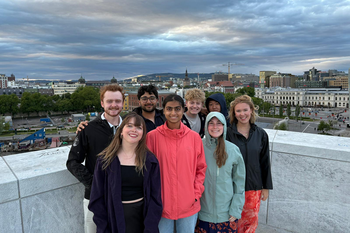 A group of 8 college students posing against a stone wall with a Norwegian city, mountains and clouds in the background.