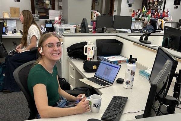 Young woman sits at a computer desk holding a coffee mug.