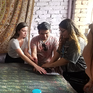 Three college students lean over a tool used for fabric printing while gathered around a piece of fabric on a table.