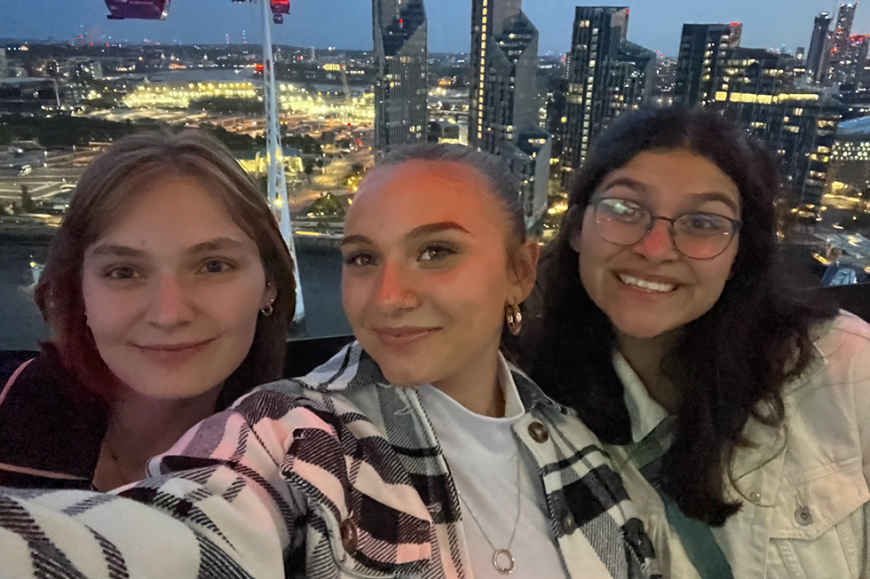 3 students pose in front of a night skyline.