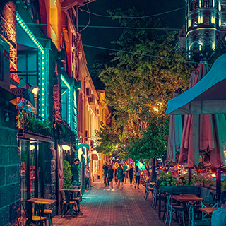 People stand on the sidewalk between brightly lit buildings at night in Yerevan, Armenia.