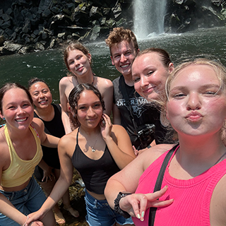 Group of college students take a photo of themselves in front of a waterfall, one flashing the peace sign.
