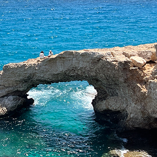 Two people sit on a rock bridge over the ocean.