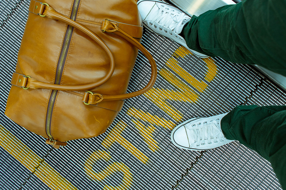 Person standing on an airport moving walkway with their leather carry-on bag. The word 'stand' appears in yellow letters across the walkway.