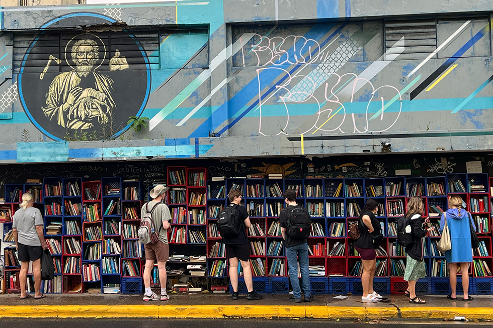 People stand in front of an outdoor wall of books made from milk crates.