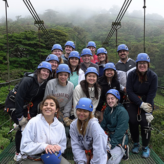 Group of young adults stand on a sky bridge wearing blue helmets, with mist and trees below and behind them.