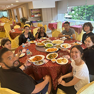 A group sits at a round table with a red tablecloth eating.