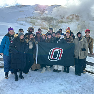 Group poses with the University of Nebraska Omaha flag in front of a snowy geyser.