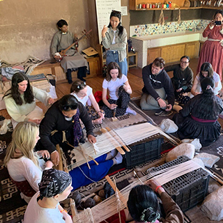 A group of people sits on the floor in a circle around a loom.