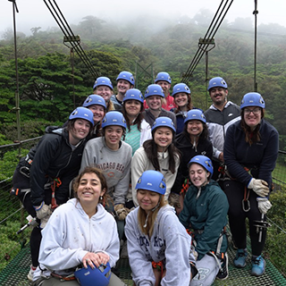 Group wearing blue helmets pose on a suspension bridge in a cloud forest.