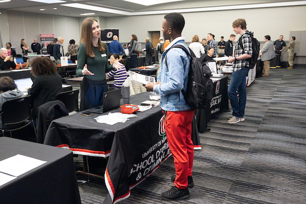 a young man of color talks to an academic advisor in a room full of tables with information about majors.