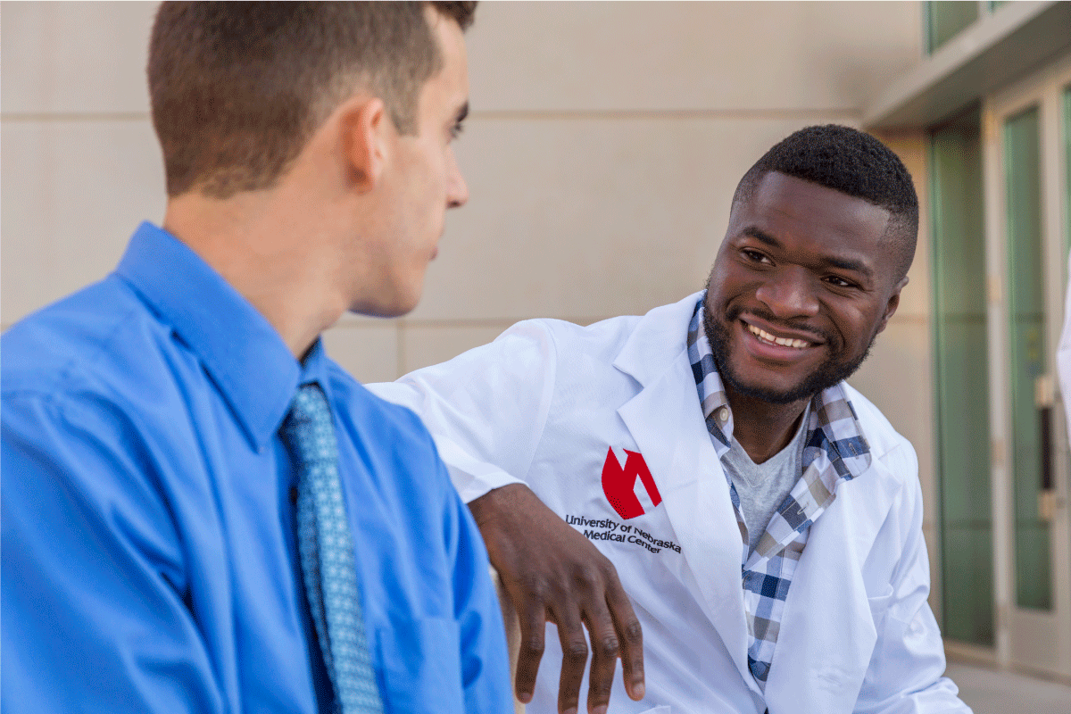 A man in a white lab coat smiles at a man in a blue shirt.