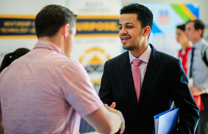 A professionally dressed student in a suit smiles and shakes hands with another person at what appears to be a career fair or networking event.
