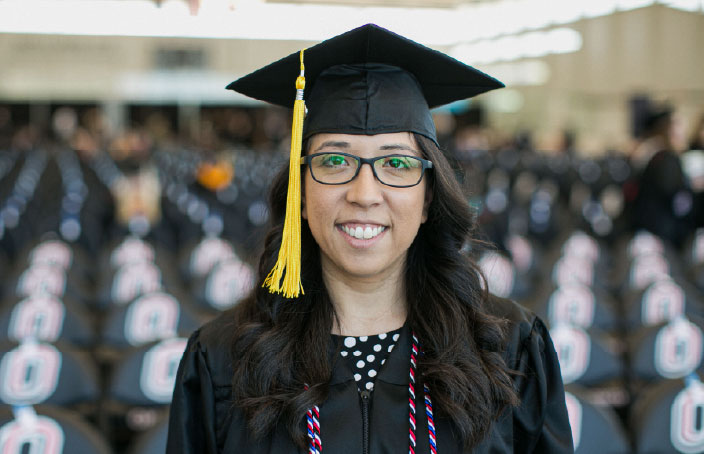 A graduate wearing a cap and gown smiles while standing in an auditorium filled with UNO-branded chairs.