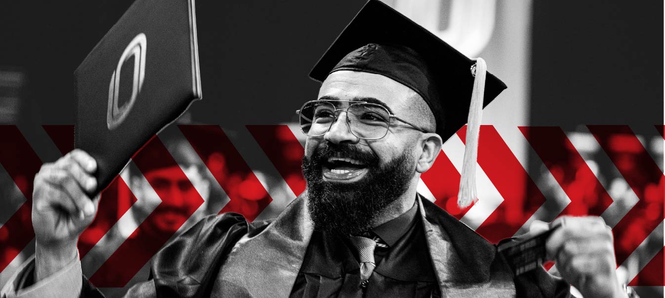 A smiling graduate in a cap and gown holds up a diploma cover during a commencement ceremony. The black-and-white image features red chevron graphics in the background.