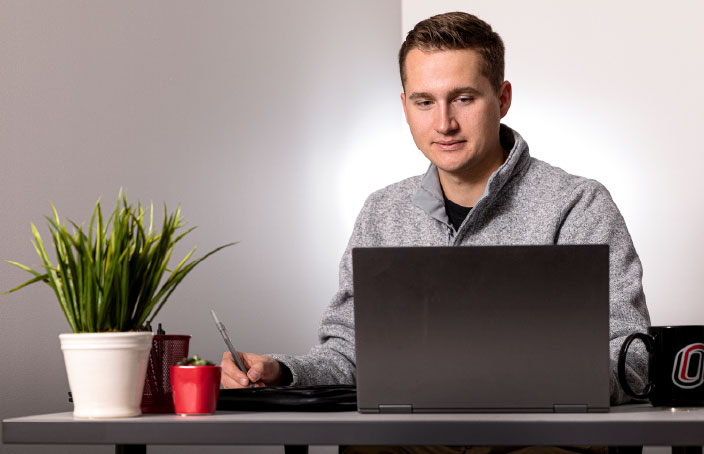 A student sits at a desk working on a laptop, with a plant, office supplies, and a UNO-branded mug nearby.