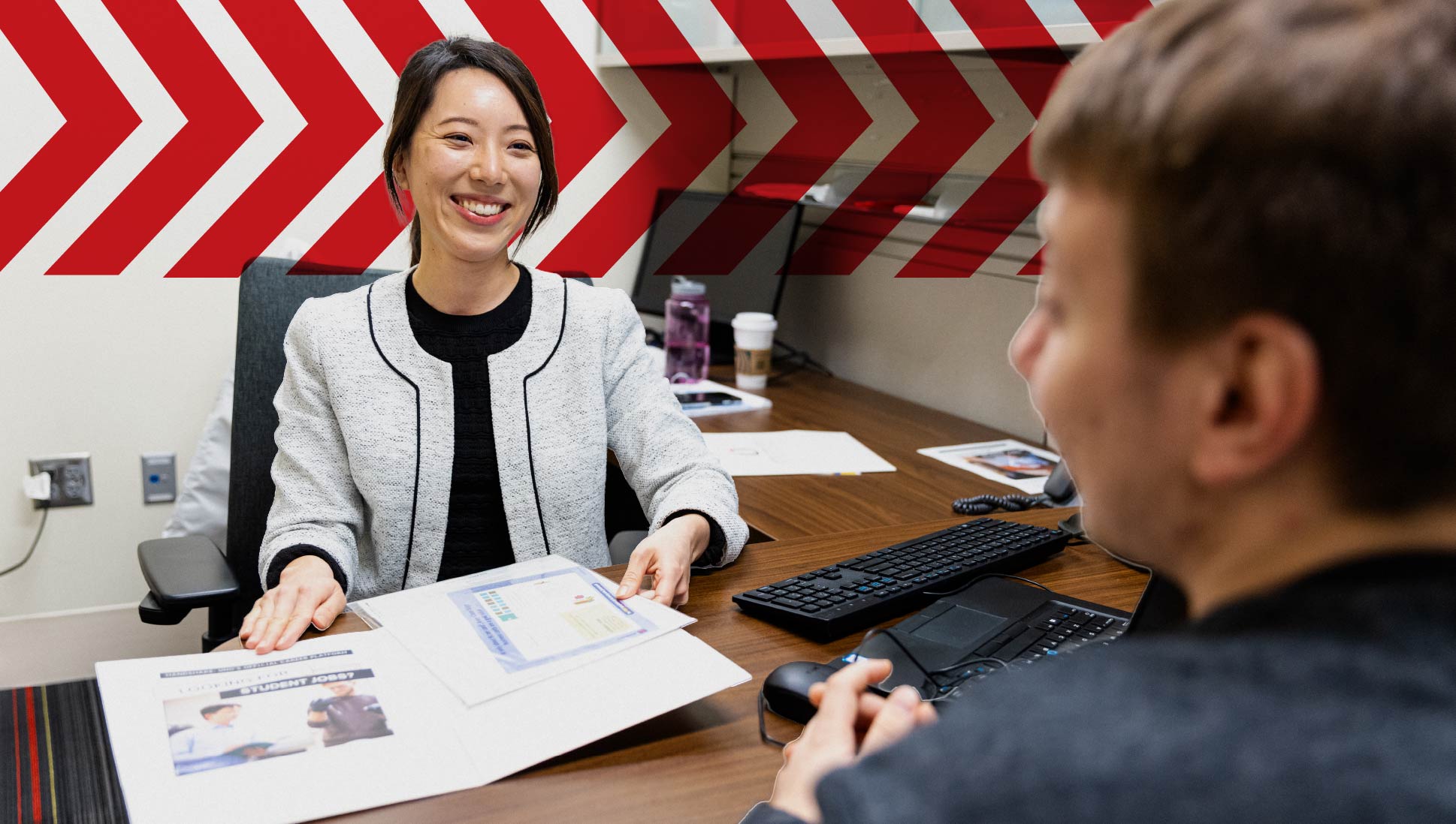 A student meets with a UNO success coach in an office, smiling while reviewing paperwork together, with red chevron graphics in the background.