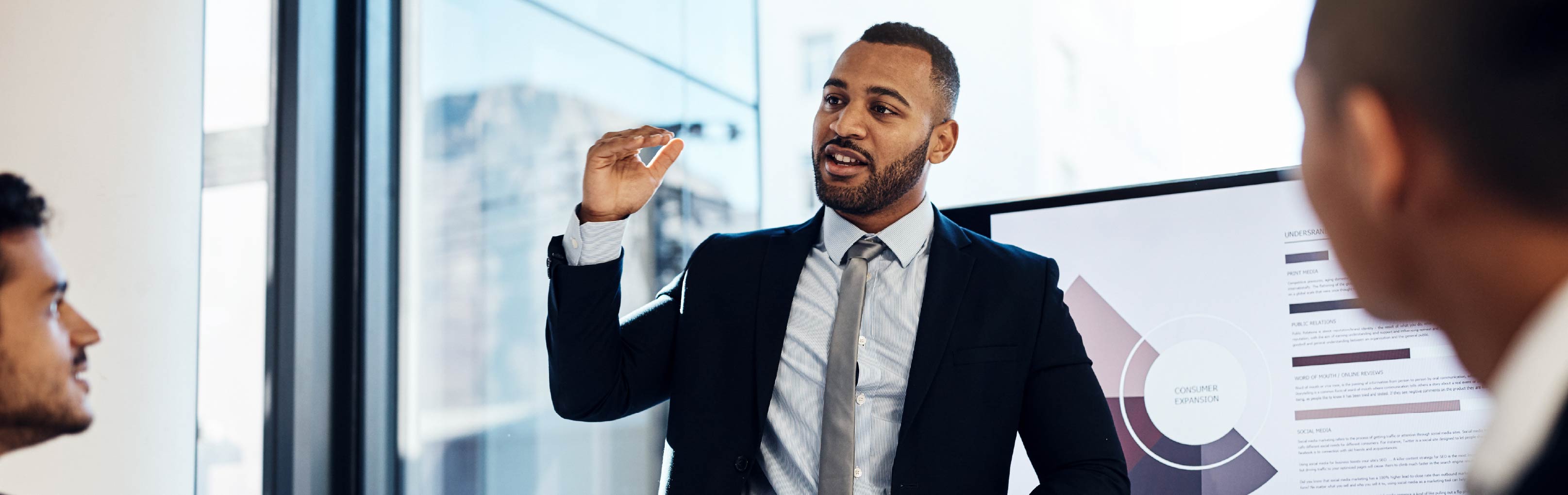 Business professional in a suit presenting to colleagues during a meeting, gesturing toward a presentation screen in a modern office conference room.