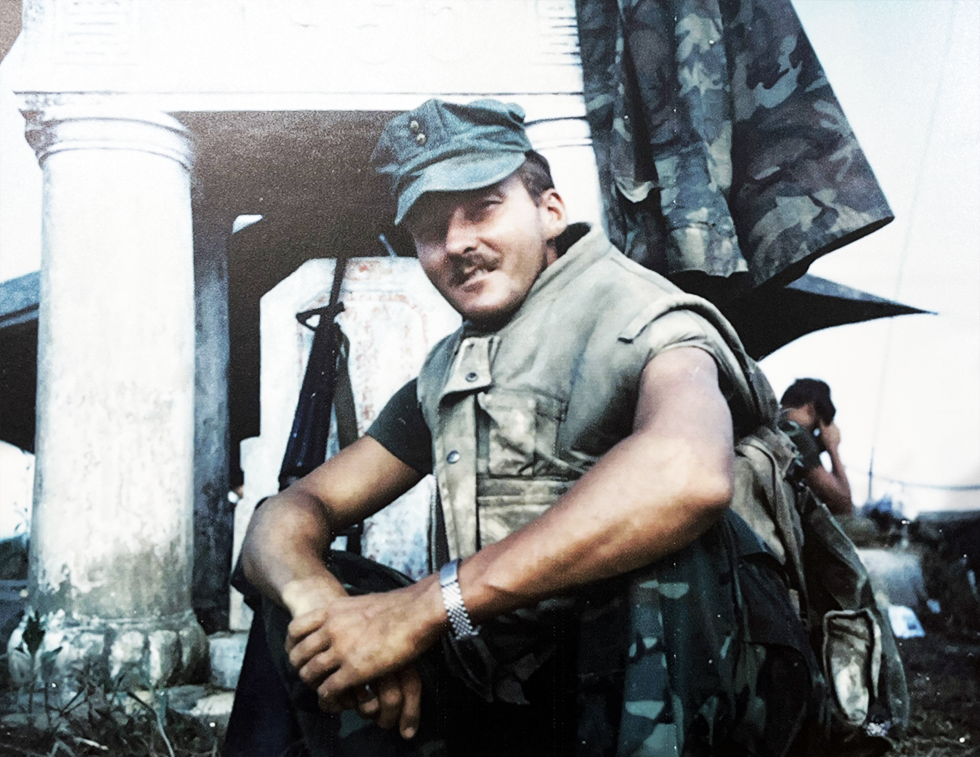 Bill Baines sitting in front of a structure in Marine uniform while deployed.