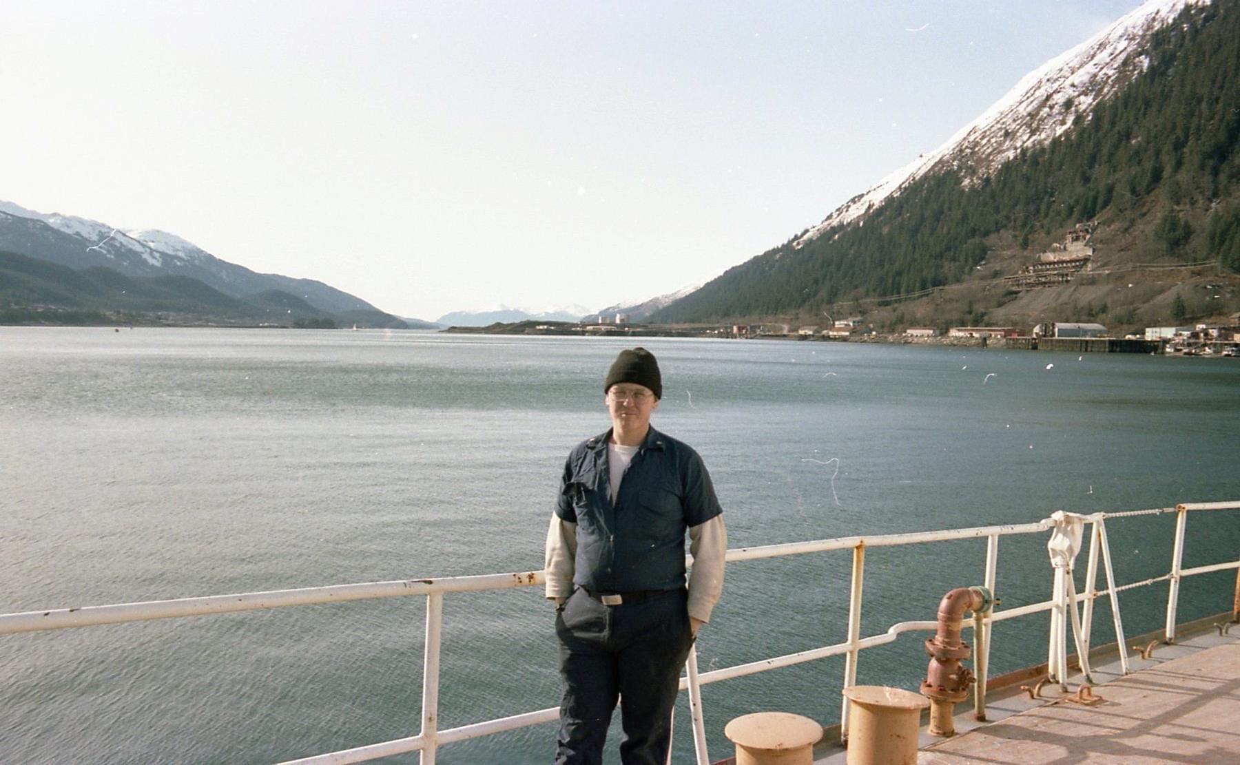 Lennie Roth in Coast Guard daily uniform on a boat while on duty with water and mountains in background.