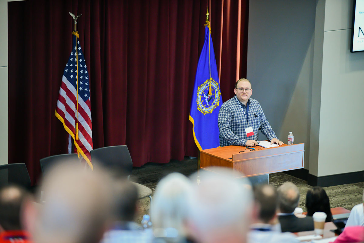 A man wearing a blue plaid shirt and glasses stands at a podium in front of an audience. 