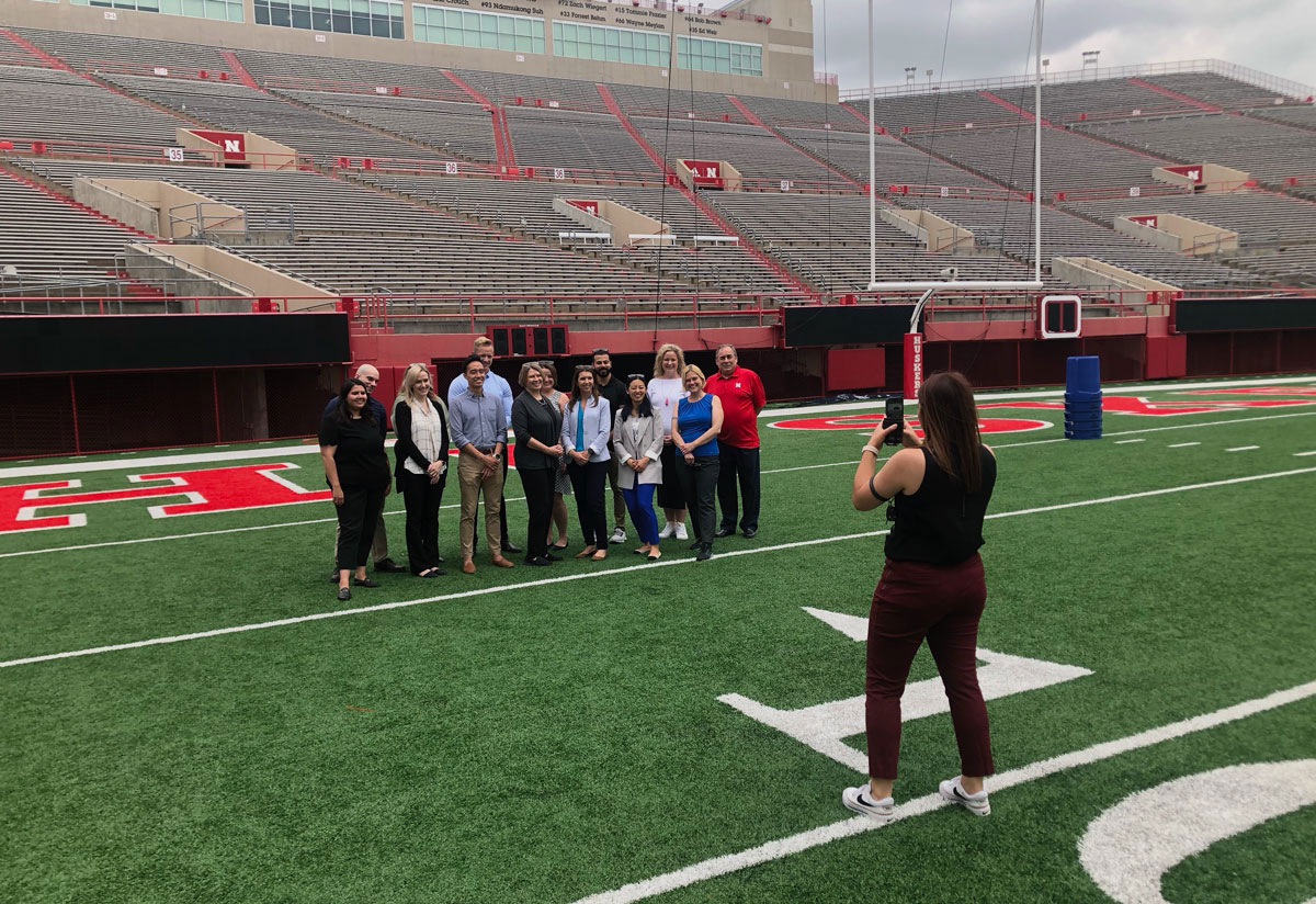 A group of about 10 people pose for a photo in the endzone of Memorial Stadium. A photographer stands in front of them. 