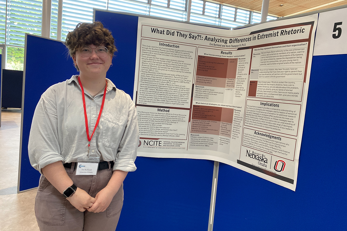 A woman with short brown hair and glasses stands next to a research poster that reads "What Did They Say?!: Analyzing Differences in Extremist Rhetoric."