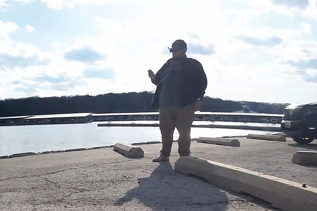 A student stands near a body of water, silhouetted in the sun. He is wearing a headset and appears to be piloting a drone. 