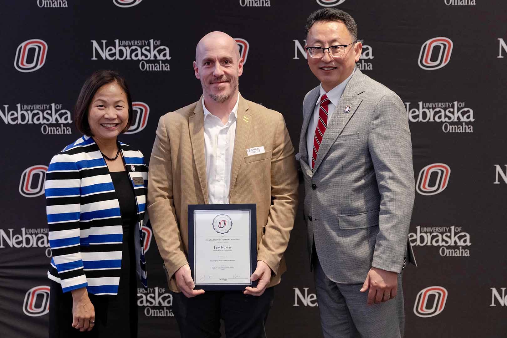 Hunter stands in front of a UNO themed background with an award in his hands. Chancellor Li stands to his left and Senior Vice Chancellor He to his right. 