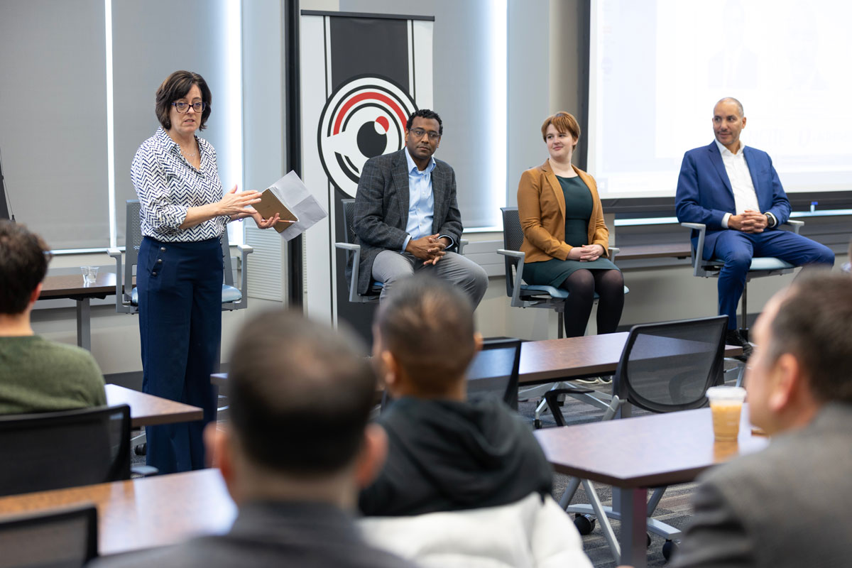 One person stands while three people sit in front of an audience in a clean, well-lit place. 