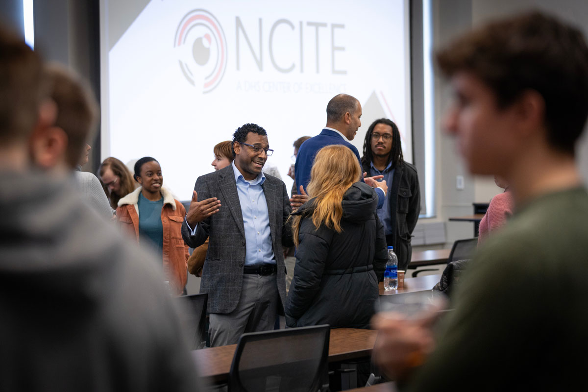 Ian Moss, wearing a gray blazer and blue shirt, speaks to a student in a black coat in front of a projected slide that reads NCITE.