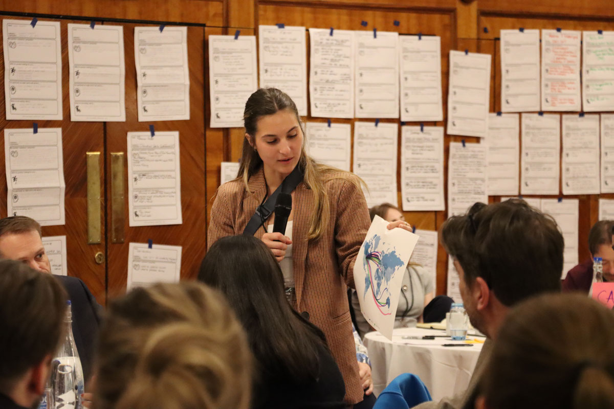 Callie Vitro speaks into a microphone at the IAPSS conference. She is holding a map with lines drawn in marker connecting various countries to each other. 