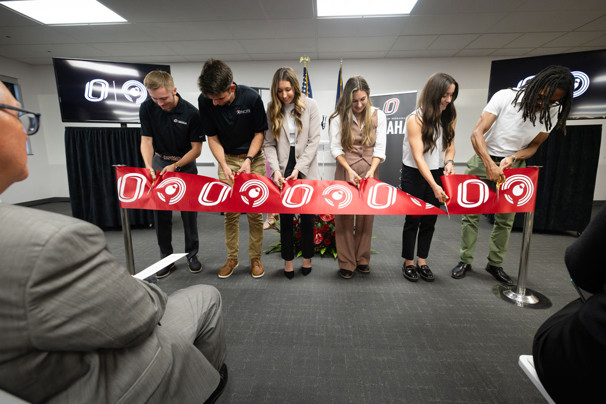 A row of six NCITE students are lined up before a red ribbon bearing the NCITE birdie and UNO O logos. They are stooped over cutting the ribbon. 