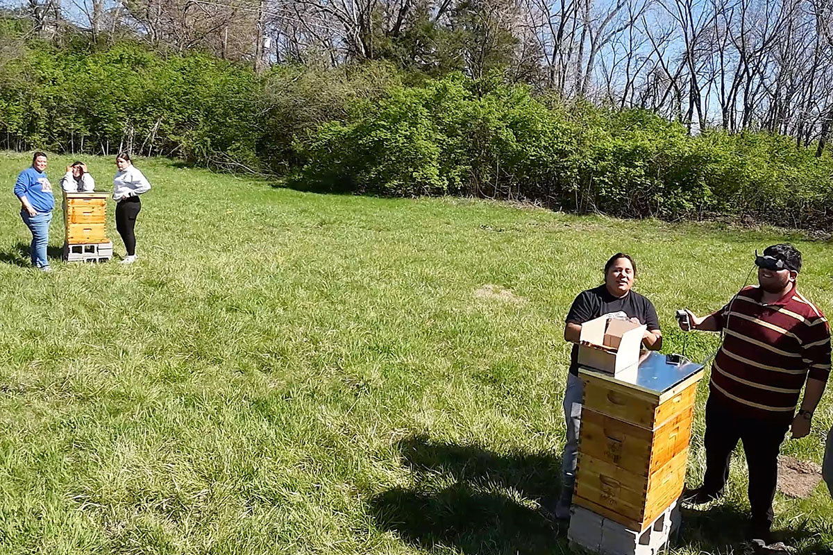 A drone shot of students standing in a grassy field. One of them is piloting the drone that's taking the photo. Others stand near stacks of wooden crates.