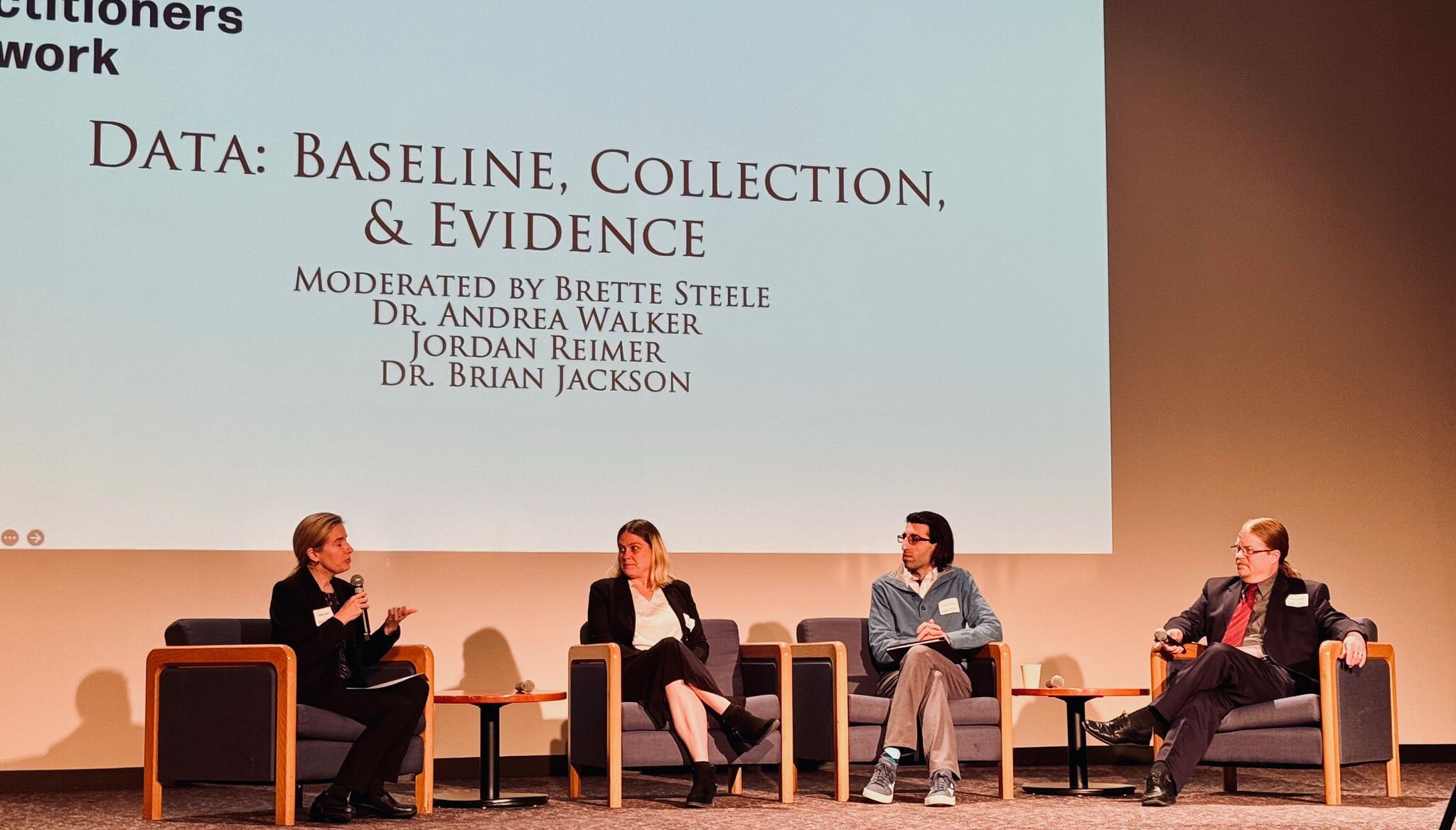 Andrea Walker sits on a stage with three people including a moderator and two other panelists. In the background is a Power Point presentation saying "Data: Baseline, Collection, & Evidence."