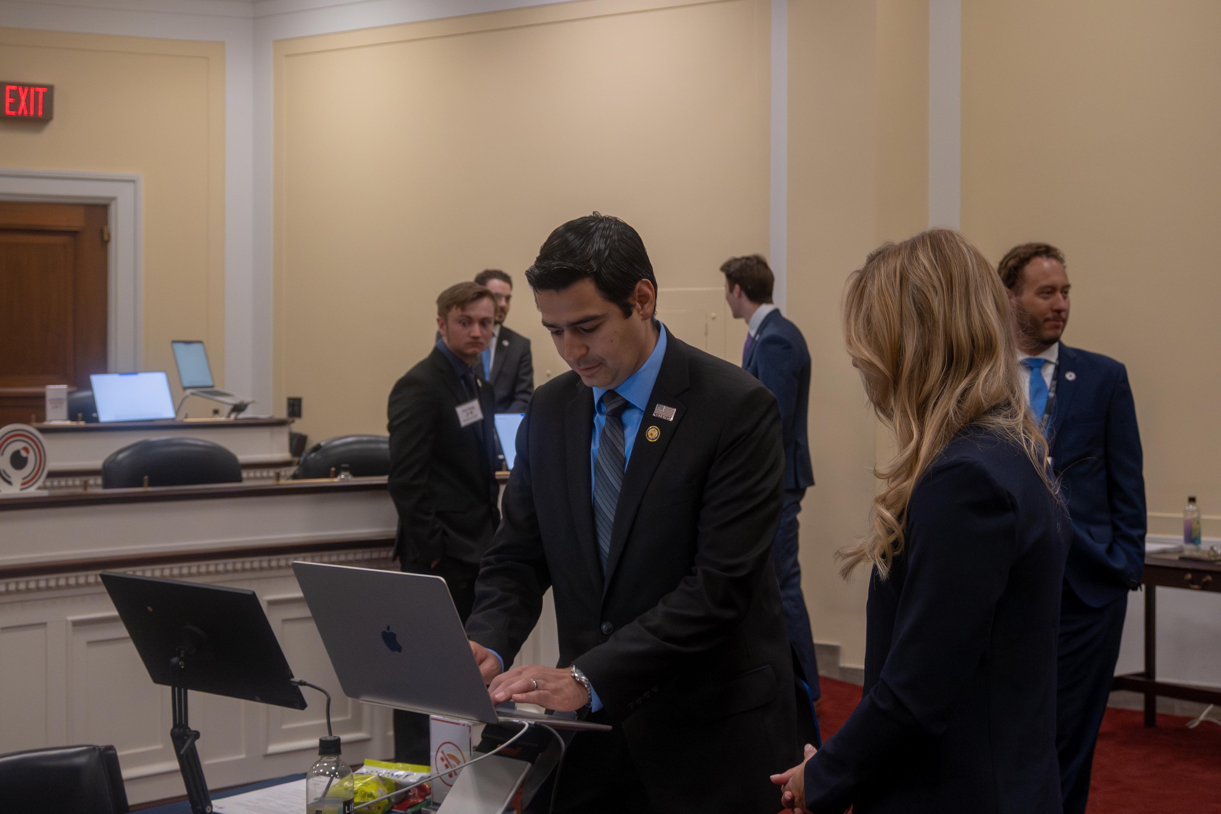 A man in a suit types on a laptop while a woman stands beside him, with other participants and laptops visible in the background.